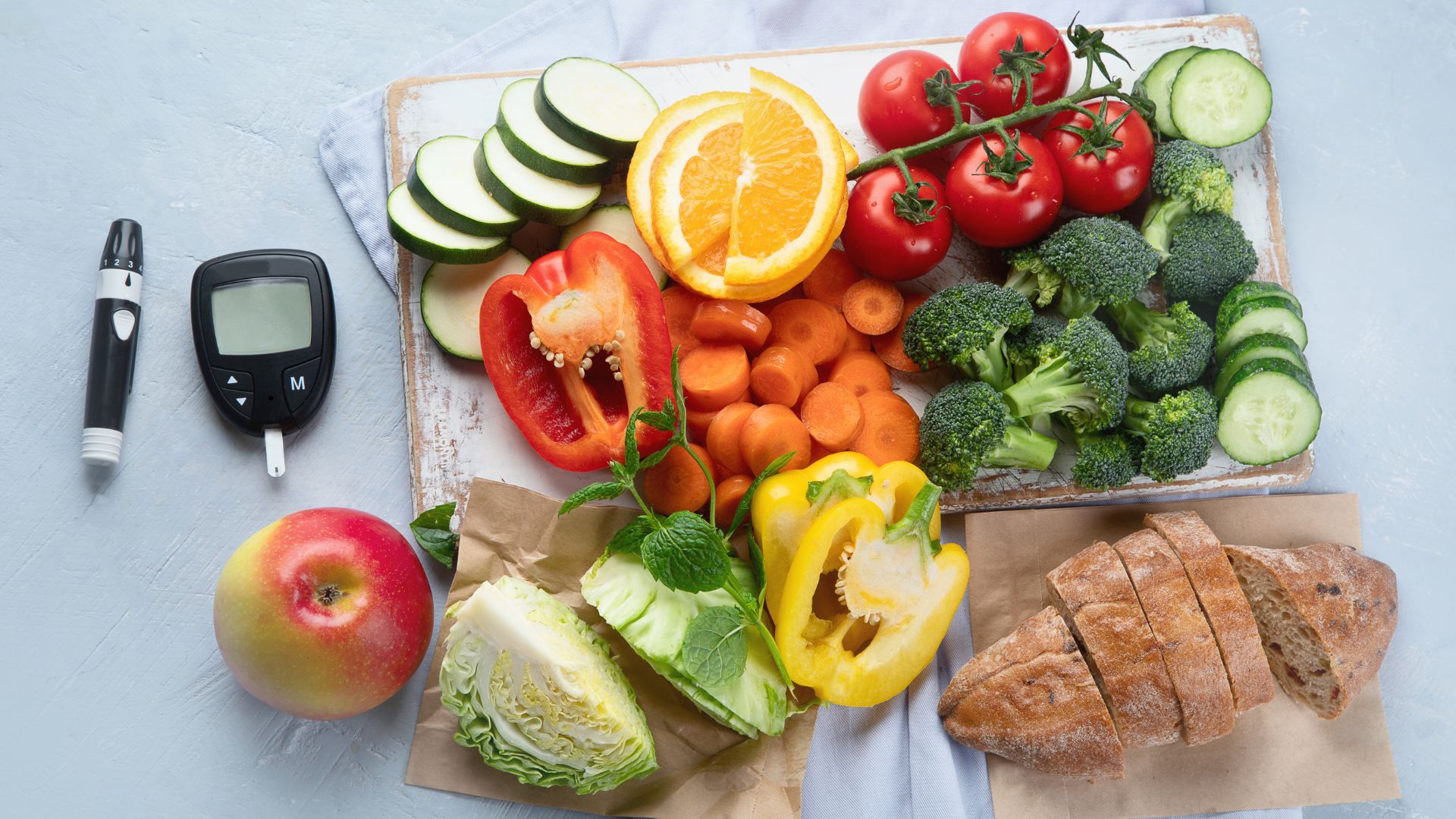 Ein Blutzuckermessgerät für Diabetiker liegt neben einem Brett, auf dem gesunde Lebensmittel wie Gemüse und Obst liegen.
