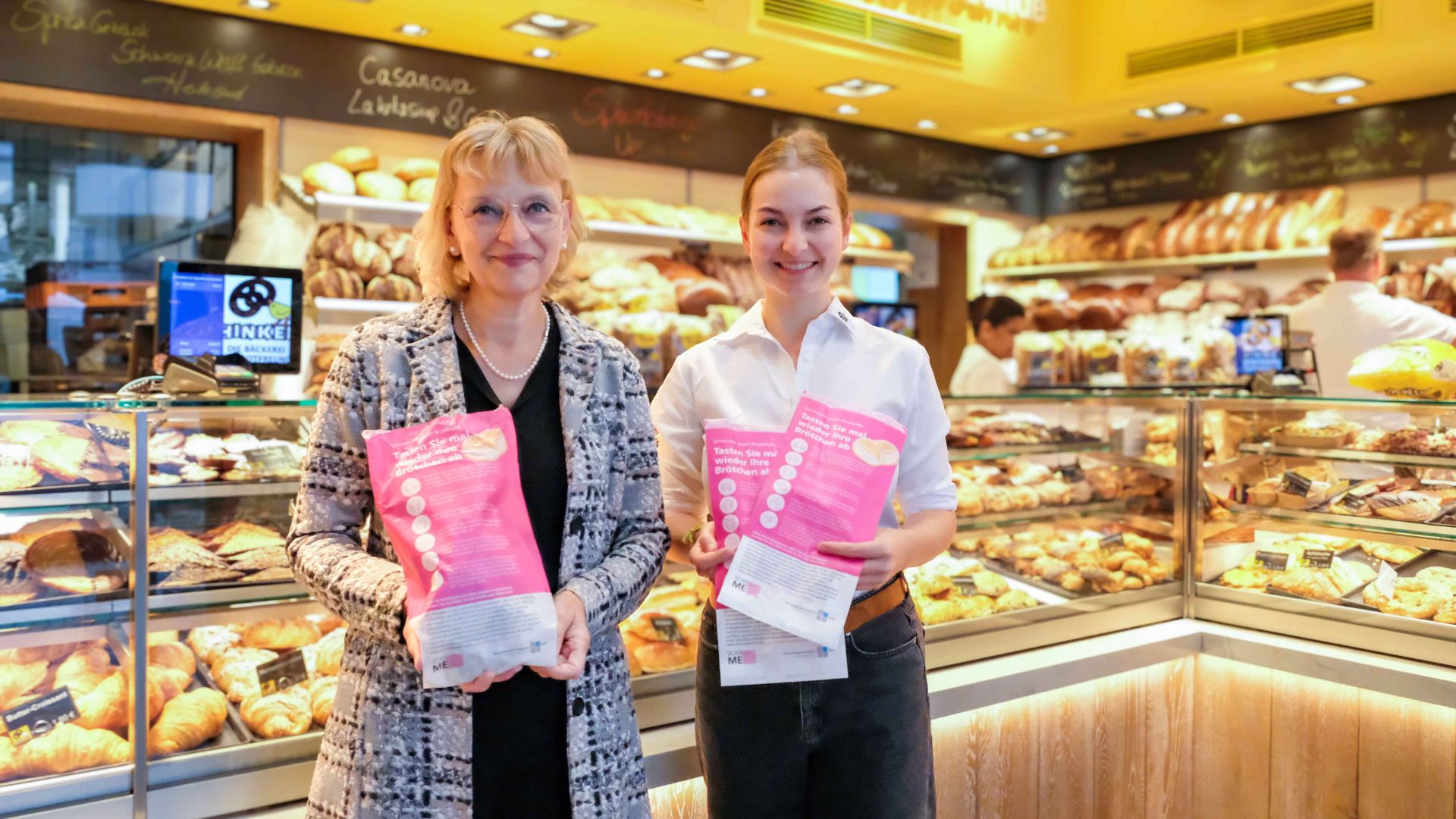 Zwei Frauen stehen in einer Bäckereifiliale und halten speziell gestaltete pinke Brötchentüten in den Händen, die Kundinnen und Kunden über Brustkrebs aufklären sollen.